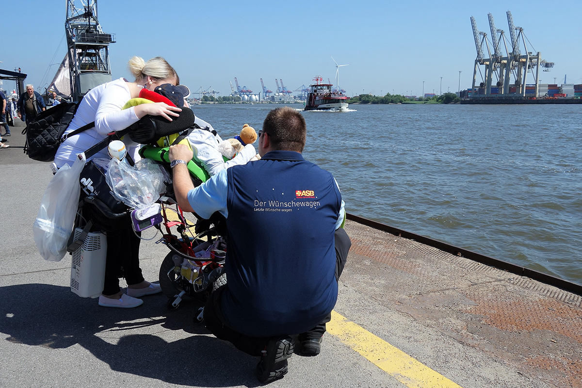 Eine Person im Rollstuhl, von hinten fotografiert, blickt bei wolkenlosem Himmel im Hamburger Hafen aufs Wasser. Neben dem Rollstuhl eine Dame und ein hockender Wünschewagen-Mitarbeiter in Dienstkleidung.