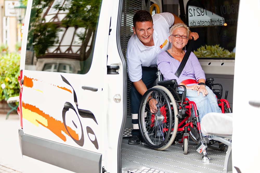 Ein Mitarbeiter des ASB Hannover kümmert sich um eine Rollstuhlfahrerin, die in einem Fahrzeug befördert wird.