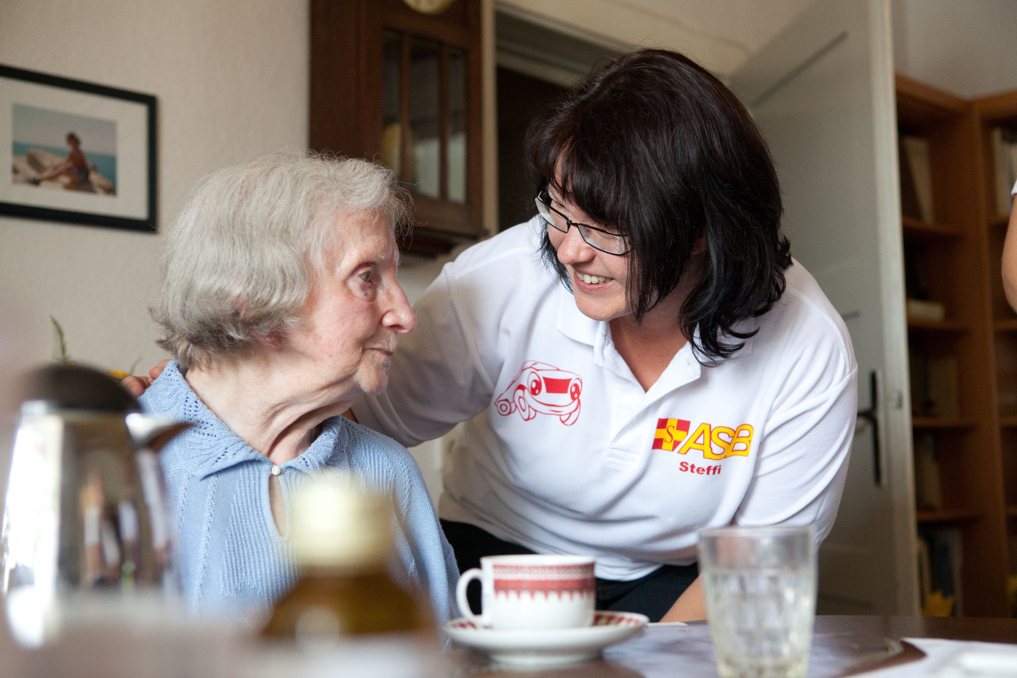 Eine ASB-Pflegerin beugt sich lächelnd zu einer Seniorin, die an ihrem Esstisch Kaffee trinkt.