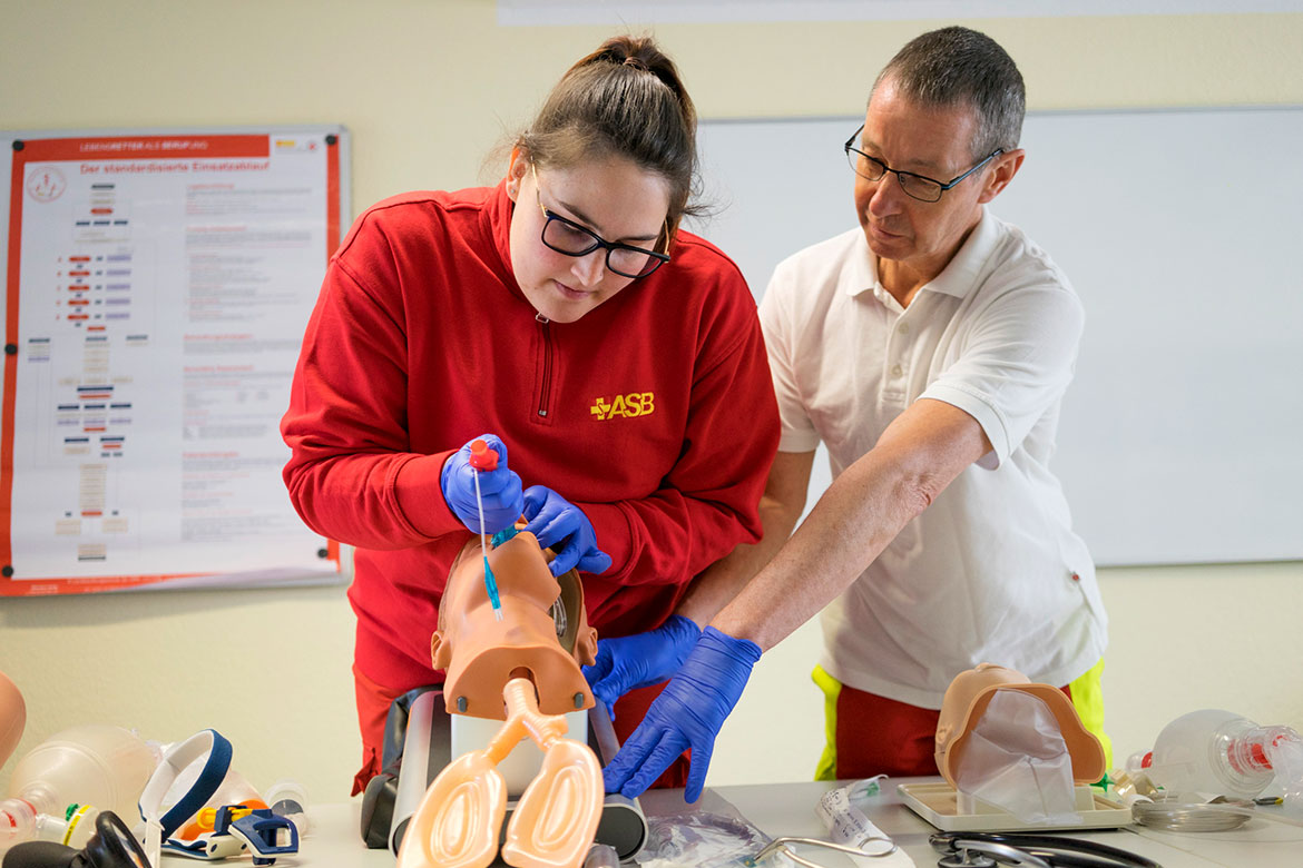 Auszubildende im Rettungsdienst schiebt einen Tubus in den Mund eines Übungsmodells. Ein Kollege unterstützt Sie dabei und hält das Modell fest.