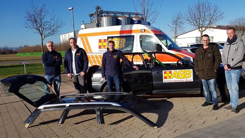 Uebungsfahrzeug-Menschenrettung1.jpg Im Vordergrund: Ein Fahrzeudach eines PKW. Dahinter stehen fünf ASB-Mitarbeiter vor dem Rest des PKW. Im Hintergrund: Ein ASB-Krankenwagen, blauer Himmel, mehrere blattlose Bäume und ein Gebäude.